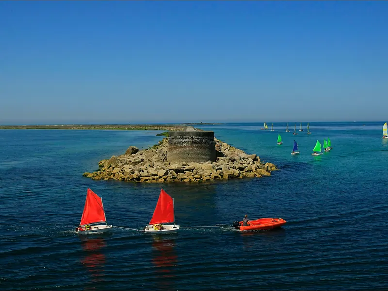 Red-sailed dinghies passing a small island fort in the Rade de Cherbourg